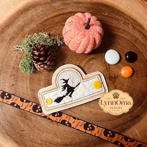 May include: A Halloween-themed design featuring a witch on a broomstick in front of a full moon, with a decorative ribbon that says "Happy Halloween". The image includes a small pumpkin, candies, and a pinecone.
