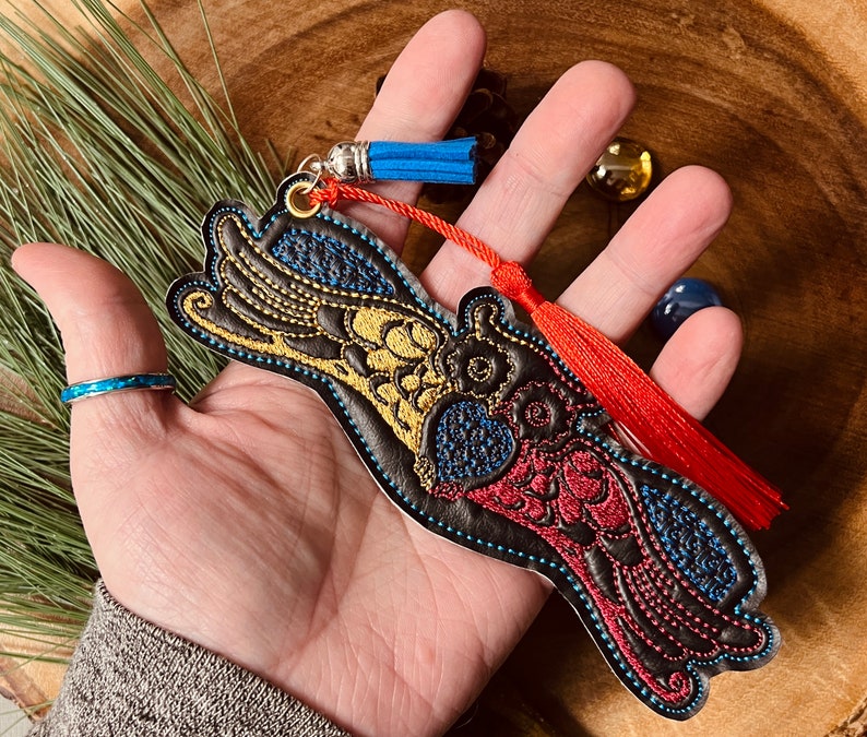 May include: A black leather bookmark with colorful embroidered bird design. The bookmark has a red and blue tassel.