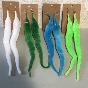 Puede incluir: Cuatro pares de pendientes con forma de gusanos peludos sujetos a tarjetas de presentación de cartón marrón. Los pendientes presentan gusanos de felpa blancos, verde oscuro, azules y verde neón con pequeños ojos móviles.