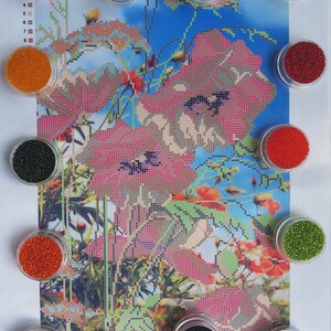May include: Assortment of small containers filled with colorful beads, used for diamond painting. The beads are in various colors, including red, orange, green, black, and white. The containers are arranged around a printed image of red poppies.