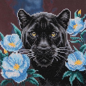 Op de afbeelding: Een kralenkunstwerk met een zwarte panter met gele ogen, omringd door blauwe en gele bloemen met groene bladeren. De achtergrond is donkerbruin.