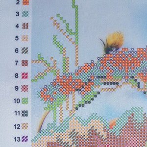 May include: A close-up of a cross-stitch pattern with a variety of colors, including orange, green, and brown. The pattern features a tree with leaves and a small bird.
