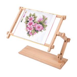 Puede incluir: Un soporte de bordado de madera con un diseño floral de punto de cruz. El soporte tiene una base rectangular y un marco ajustable que sostiene la tela. El diseño de punto de cruz muestra flores rosas y hojas verdes.