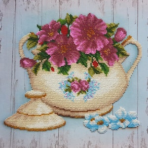 Può includere: Una teiera in ceramica bianca con un design floreale, decorata con fiori rosa e foglie verdi. La teiera ha un coperchio e un manico, ed è appoggiata su una superficie di legno.
