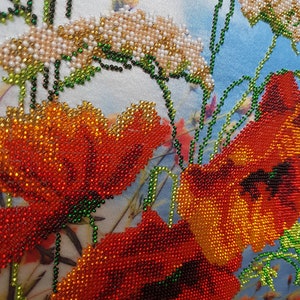 May include: Close-up of a beaded embroidery design featuring red poppies with green stems and leaves. The background is a light blue fabric with white flowers.