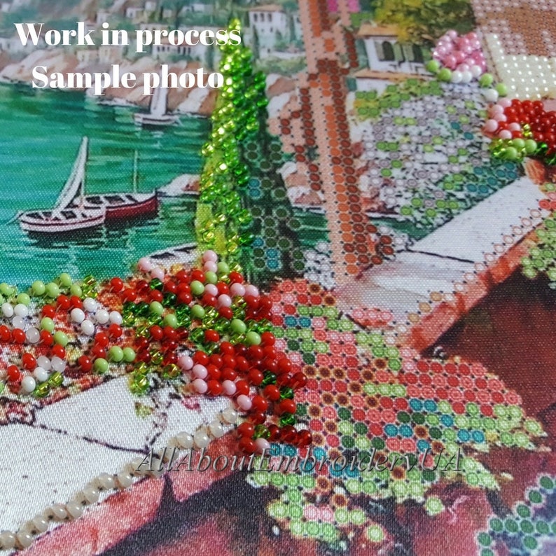 Puede incluir: Primer plano de una pintura de diamantes parcialmente terminada que representa un paisaje urbano italiano colorido con un barco en el agua. La pintura est&aacute; hecha en un lienzo con un patr&oacute;n de cuadr&iacute;cula y presenta cuentas de varios colores, incluidos rojo, verde, blanco y rosa.