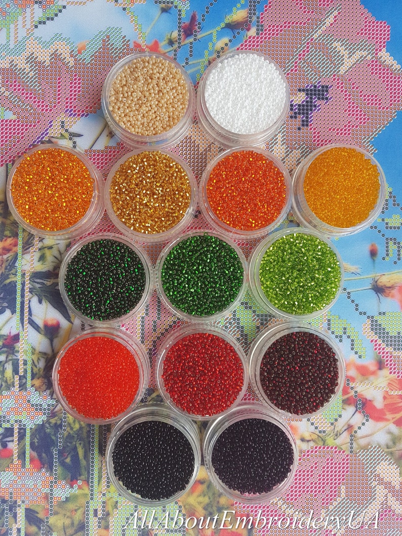 May include: Twelve small containers of seed beads in various colors, including white, gold, orange, green, red, and black. The containers are arranged in a circle on a floral patterned surface.