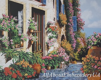 May include: A painting of a narrow, cobblestone street in a European village. The street is lined with buildings with windows and doors, and there are many potted plants and flowers in bloom. The sky is blue and there are clouds in the distance.