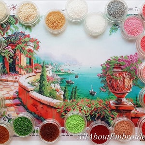 Puede incluir: Surtido de peque&ntilde;os recipientes llenos de cuentas de colores, utilizados para la pintura de diamantes. Las cuentas est&aacute;n dispuestas en c&iacute;rculo alrededor de una imagen impresa de un paisaje mediterr&aacute;neo con un edificio de techo rojo, un camino de piedra y un gran jarr&oacute;n con flores rosas.