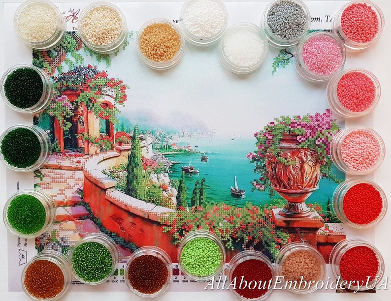 Puede incluir: Surtido de peque&ntilde;os recipientes llenos de cuentas de colores, utilizados para la pintura de diamantes. Las cuentas est&aacute;n dispuestas en c&iacute;rculo alrededor de una imagen impresa de un paisaje mediterr&aacute;neo con un edificio de techo rojo, un camino de piedra y un gran jarr&oacute;n con flores rosas.