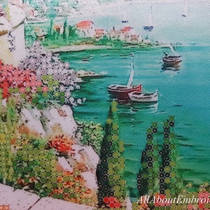 Puede incluir: Un dise&ntilde;o de bordado de punto de cruz que representa una escena costera con un mar azul turquesa, dos veleros y una costa rocosa con vegetaci&oacute;n verde y flores rojas.