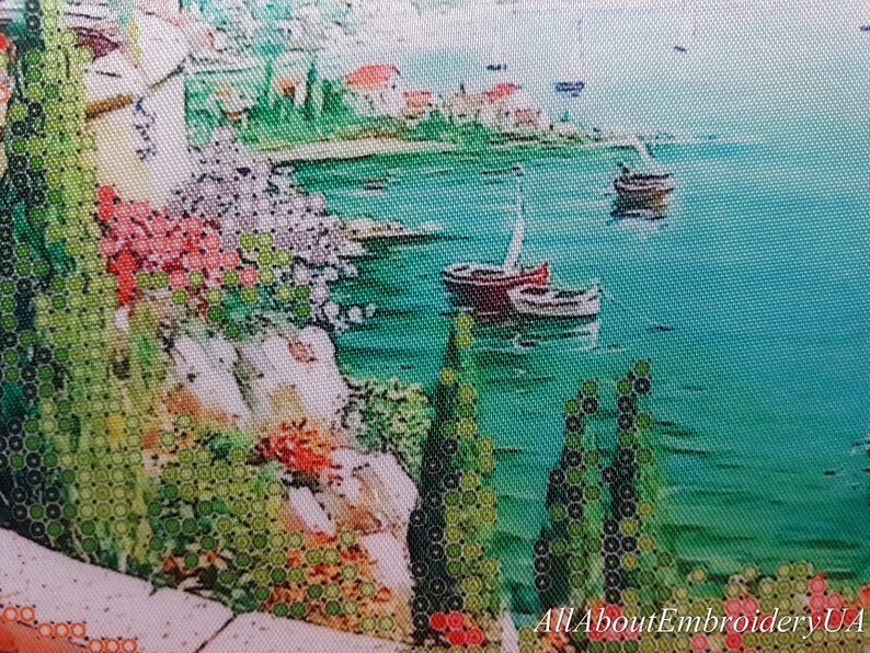 Puede incluir: Un dise&ntilde;o de bordado de punto de cruz que representa una escena costera con un mar azul turquesa, dos veleros y una costa rocosa con vegetaci&oacute;n verde y flores rojas.