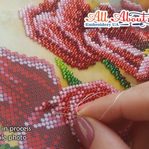 May include: Close-up of a hand beading a red and pink flower design on a canvas. The beads are small and round, and the design is intricate. The text "Work in process Sample photo" is visible in the lower left corner.