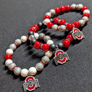Peut inclure: Trois bracelets avec des perles rouges, blanches et grises. Chaque bracelet a un charme argenté avec le logo de l'Ohio State.