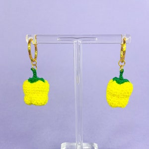 May include: A pair of yellow crocheted bell pepper earrings hanging from gold hoop earrings on a clear display stand.