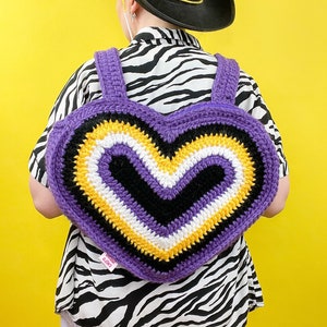 May include: A purple, black, white and yellow crocheted heart-shaped backpack. The backpack has adjustable straps and is being worn by a person wearing a zebra print shirt.