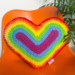 A heart shaped cushion in rainbow colours lying on an orange desk chair next to a plant.
