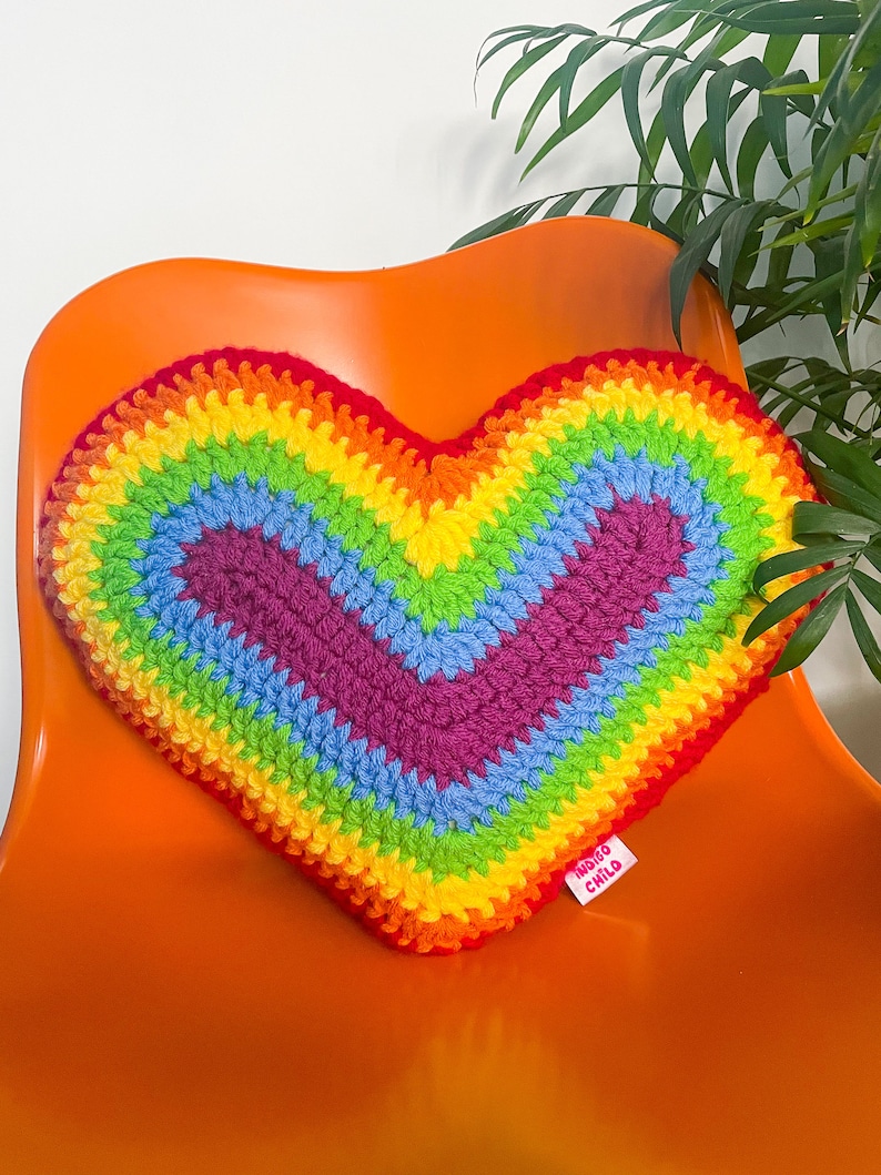A heart shaped cushion in rainbow colours lying on an orange desk chair next to a plant.