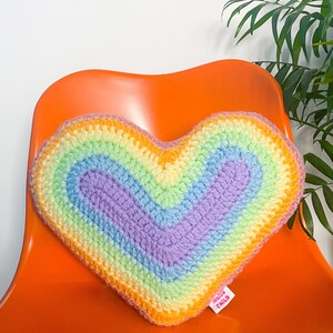 Close view of a heart shaped pillow with a pastel rainbow design sat on a desk chair in front of a white wall.