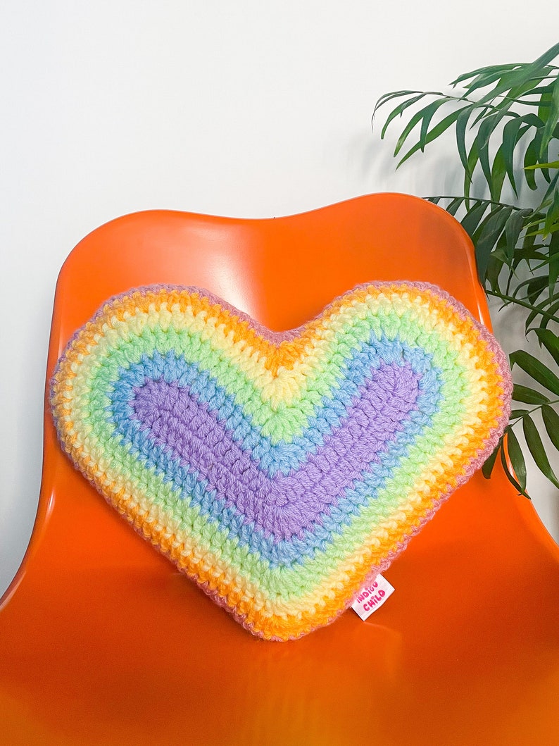 Close view of a heart shaped pillow with a pastel rainbow design sat on a desk chair in front of a white wall.