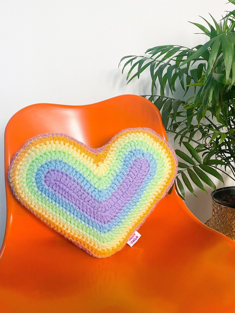 A heart pillow in pastel rainbow colours sat upright on an orange desk chair next to a plant.
