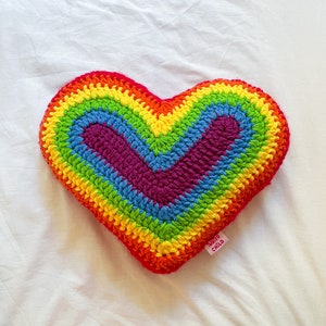 Flat lay of a crochet cushion shaped like a heart in rainbow colours lying on a white sheet.