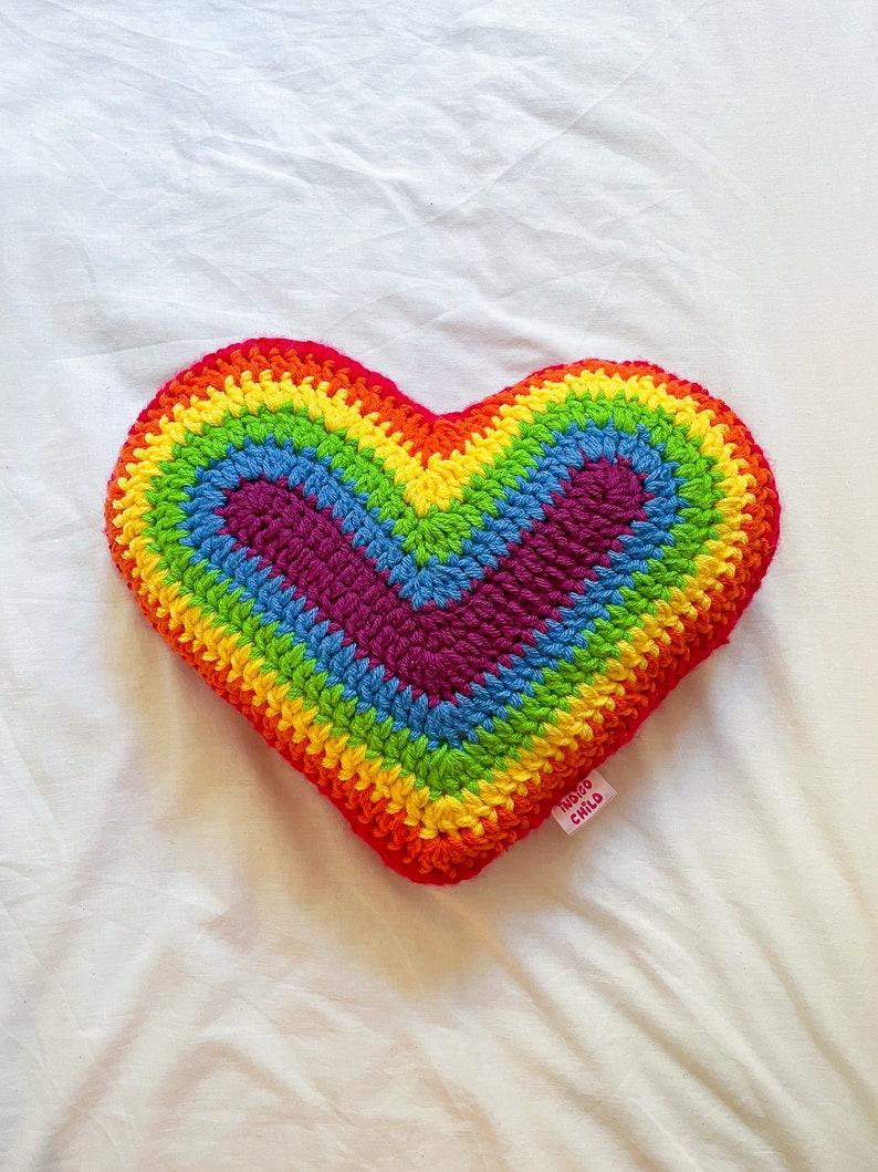 Flat lay of a crochet cushion shaped like a heart in rainbow colours lying on a white sheet.
