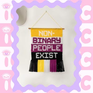 May include: A crocheted wall hanging with the words "NON-BINARY PEOPLE EXIST" in yellow, white, purple, and black yarn. The hanging has fringe on the bottom and is labelled "INTERMEDIATE".