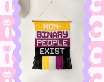 May include: A crocheted wall hanging with the words "NON-BINARY PEOPLE EXIST" in yellow, white, purple, and black yarn. The hanging has fringe on the bottom and is labelled "INTERMEDIATE".