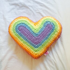 Flat lay of a pastel rainbow cushion shaped like a heart lying on a white sheet.