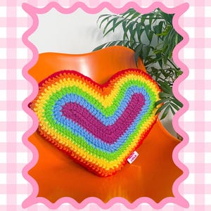 Pattern cover of a crochet pillow shaped like a heart in rainbow colours sat on an orange desk chair with the words rainbow heart cushion pillow and skill level intermediate.