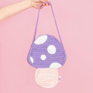 May include: A whimsical, handmade crochet mushroom-shaped bag in shades of purple, white, and pink. The bag features a purple top with white polka dots, a pink stem, and a braided purple strap. The bag is held up by a hand.
