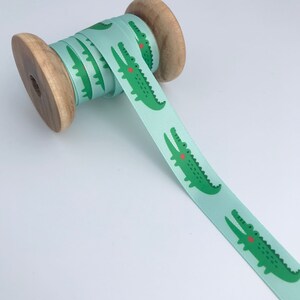 May include: A spool of light blue satin ribbon with a repeating pattern of green crocodiles. The crocodiles have red eyes and are facing to the right.