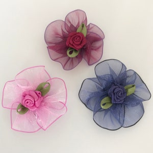 May include: Three decorative flower hair accessories. Each flower has a sheer, petal-like design in shades of pink, purple, and blue. A small rose and green leaves are at the centre of each flower. The accessories are on a white background.