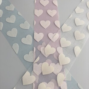 May include: Three rolls of white tulle ribbon with a white heart pattern. The ribbon is layered on a light gray background.