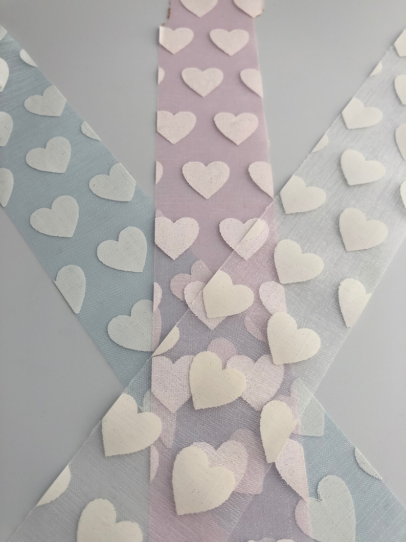 May include: Three rolls of white tulle ribbon with a white heart pattern. The ribbon is layered on a light gray background.