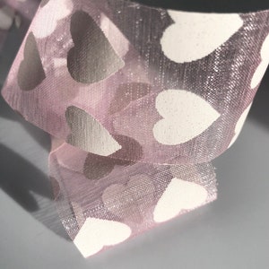 May include: Pink sheer ribbon with white heart-shaped patterns. The ribbon is folded and the hearts are visible on both sides.