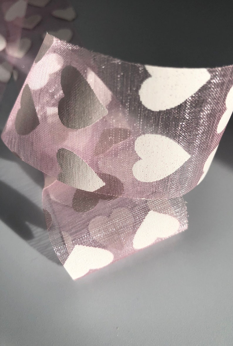 May include: Pink sheer ribbon with white heart-shaped patterns. The ribbon is folded and the hearts are visible on both sides.