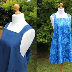 May include: Two aprons displayed on dress forms. One is solid navy blue with a square neckline and a pocket. The other is a bright blue with a pattern of seashells and fish. Both aprons have wide shoulder straps.
