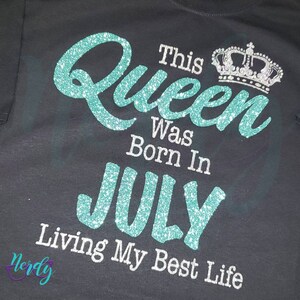Puede incluir: Una camiseta negra con un dise&ntilde;o de purpurina turquesa que dice "This Queen Was Born In July Living My Best Life". Una corona de purpurina plateada est&aacute; por encima de la palabra "Queen".