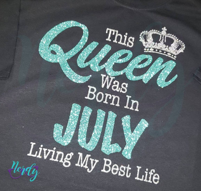 Puede incluir: Una camiseta negra con un dise&ntilde;o de purpurina turquesa que dice "This Queen Was Born In July Living My Best Life". Una corona de purpurina plateada est&aacute; por encima de la palabra "Queen".