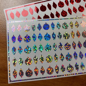 May include: Two sheets of sticker paper with a variety of Christmas ornament designs. The top sheet features red glitter ornaments and the bottom sheet features iridescent glitter ornaments.