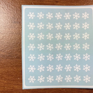 May include: A sheet of white snowflake stickers on a light blue background. The sheet is 6 inches by 6 inches and contains 49 snowflakes.
