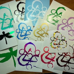 May include: Assortment of stickers featuring a stylized logo with a variety of colors and finishes, including holographic, glitter, and metallic. The logo is a stylized design of three interlocking circles.