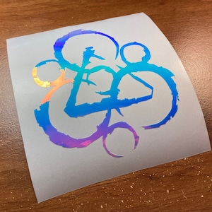 May include: A holographic vinyl sticker with a blue, purple, and gold design. The design is a stylized symbol with multiple circles and a triangle.