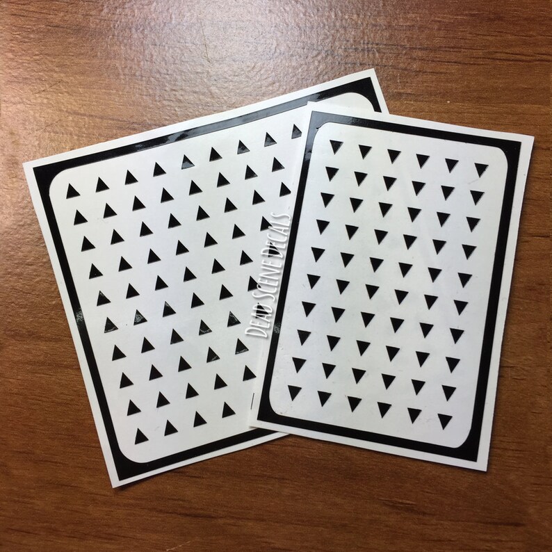 Pu&ograve; includere: Due fogli di adesivi neri e bianchi con un motivo ripetitivo di piccoli triangoli neri su sfondo bianco. I fogli di adesivi sono etichettati "Dead Scene Decals".