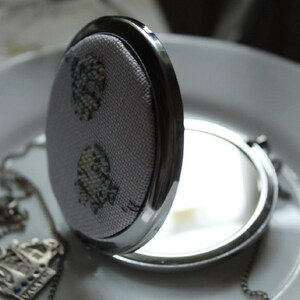 May include: A silver compact mirror with a floral design on the lid. The mirror is open and the reflection is visible.