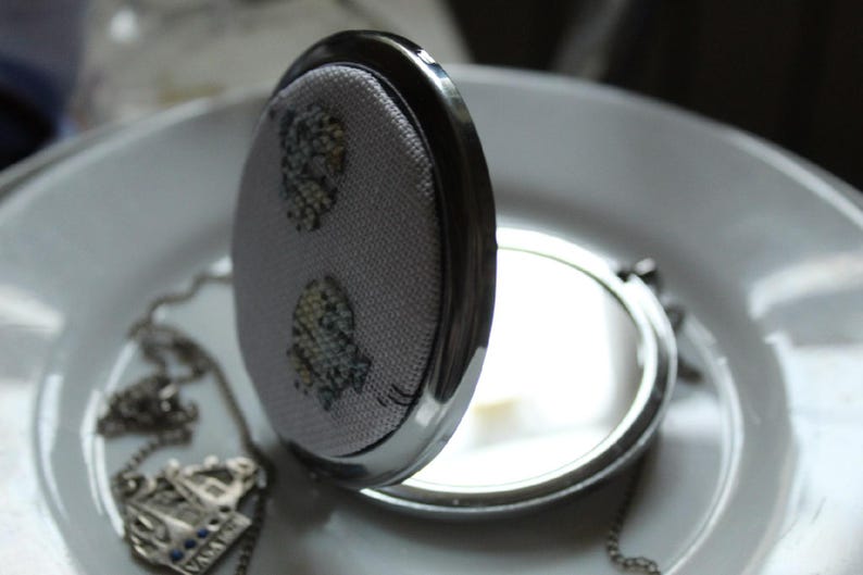 May include: A silver compact mirror with a floral design on the lid. The mirror is open and the reflection is visible.