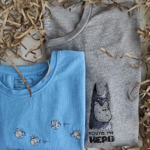 May include: Two t-shirts, one light blue with a design of four cartoon fish and the other gray with a cartoon Batman design and the text "You're my hero".