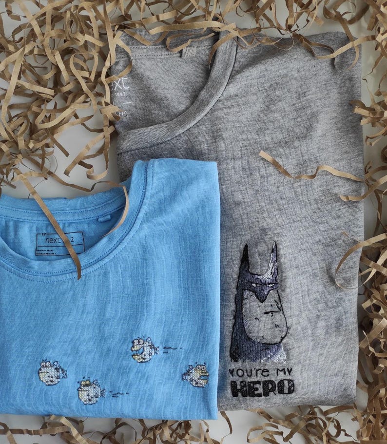 May include: Two t-shirts, one light blue with a design of four cartoon fish and the other gray with a cartoon Batman design and the text "You're my hero".